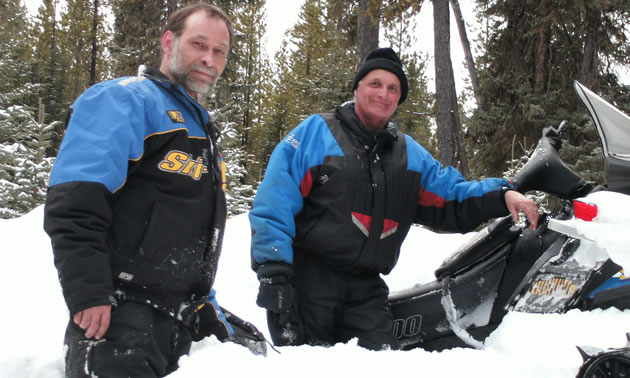 Photo of a son and his dad standing in the snow beside a snowmobile that is stuck. 