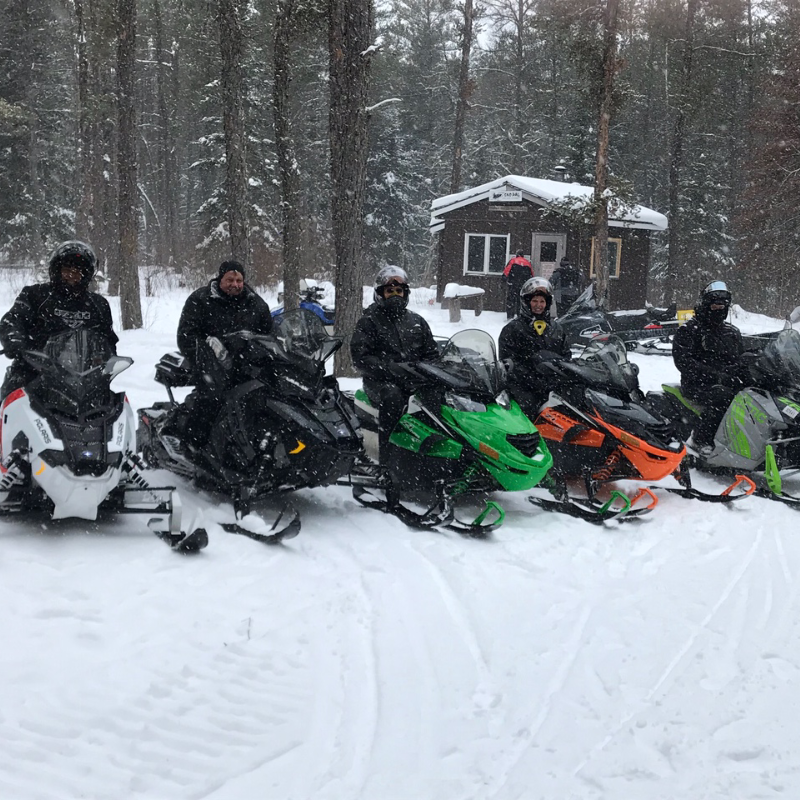 Five snowmobilers pose for a photo in front of a warm-up shelter as snow falls.