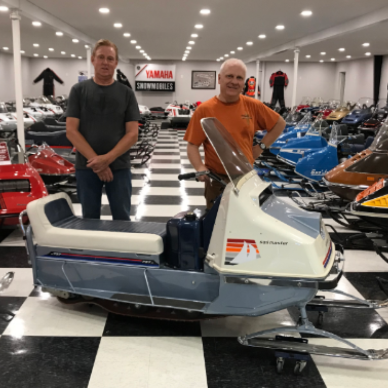 Two men stand behind a 1966 prototype Evinrude Sail Master in a room full of vintage snowmobiles