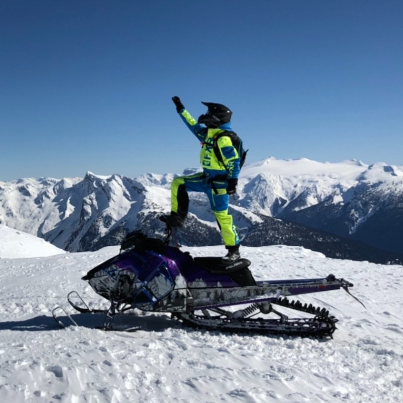 Andrew McKenzie stands on top of his black snowmobile, holding up his hand with a skyline of mountains in the distance. 