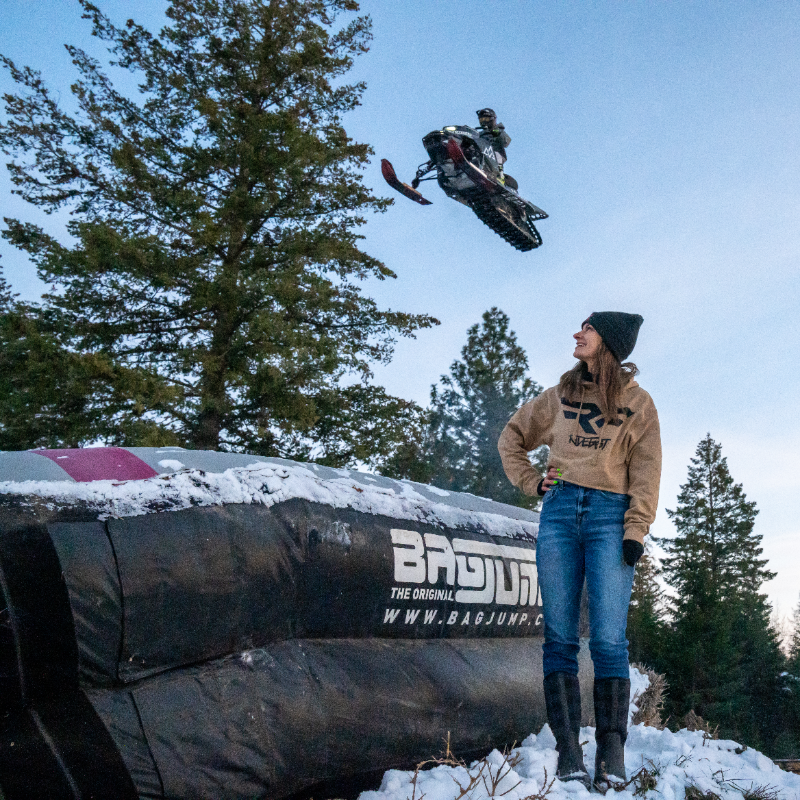 Brandon Milford soars into the air on his snowmobile while a woman looks on next to an inflatable landing platform. 