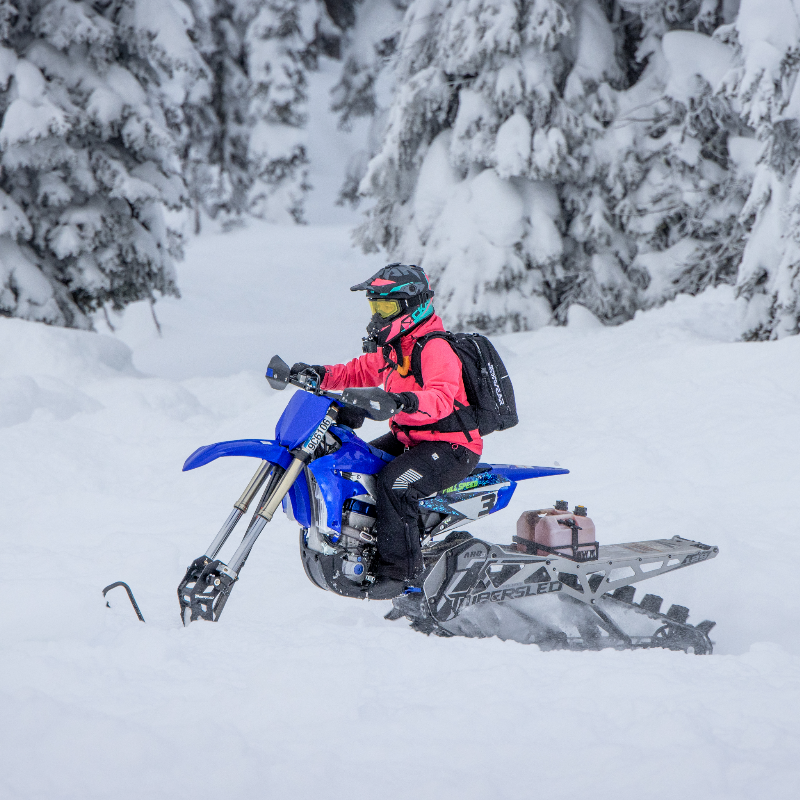 Cassandra Mainville wears a pink jacket and sits on a blue snow bike in a snowy forest. 