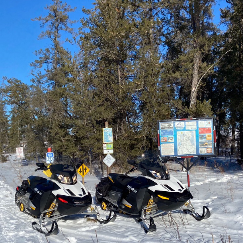 Two snowmobiles are parked in front of road signage next to a groomed trail.