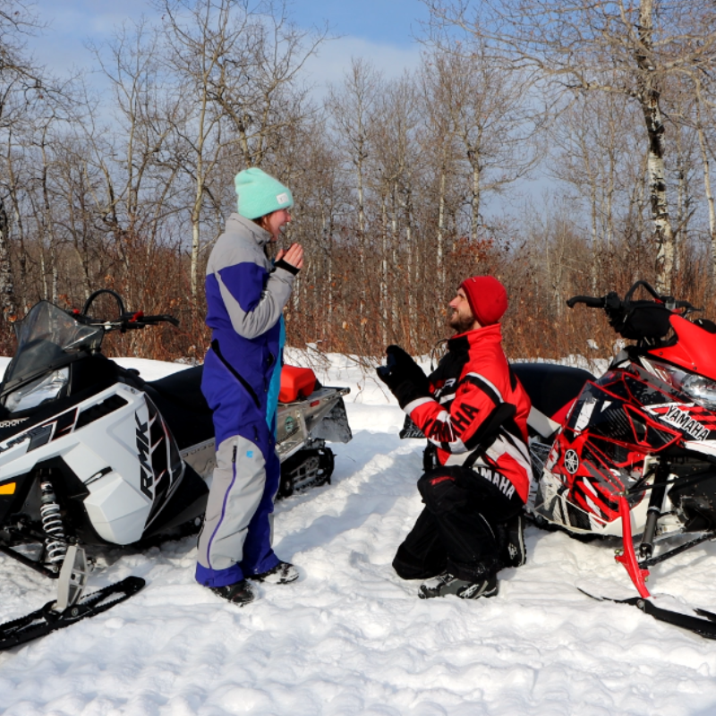 A man wearing red and black proposes to a woman wearing blue on a snowy trail next to snowmobiles. 