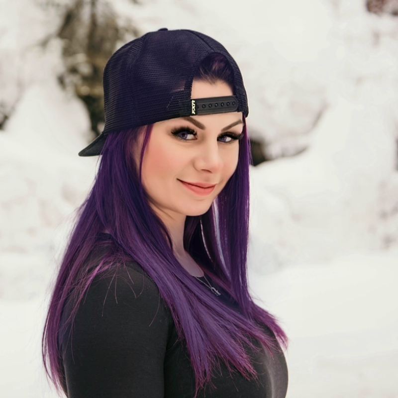 Kelsey Fay has long purple hair and wears a backwards baseball cap and black shirt. She smiles while standing in the snow. 