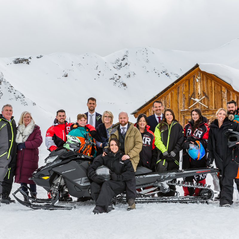 At Toby Creek Lodge, a group of wedding guests pose behind a black snowmobile. 