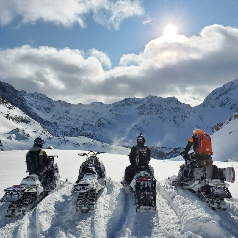 A row of snowmobilers parks at the top of a snowy mountain as the sun breaks through the clouds up above. 