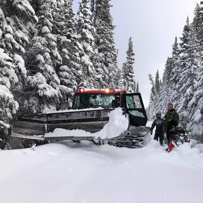 A snowmobile trail groomer is parked next to two men on a snowmobile trail. 