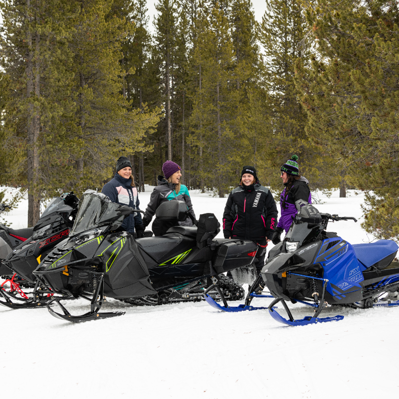 Four women visit and laugh while taking a break on their snowmobiles. 