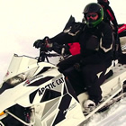Erin, wearing a black and red snowsuit, sits on his new white arctic snowmobile while a fellow rider looks on from behind on his own sled.
