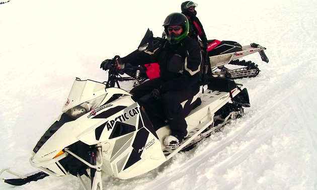 Erin, wearing a black and red snowsuit, sits on his new white arctic snowmobile while a fellow rider looks on from behind on his own sled.