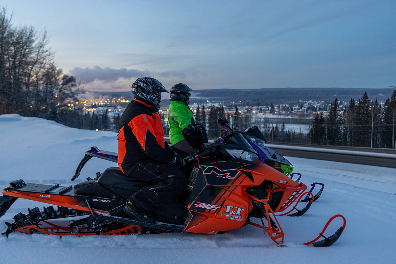 Two snowmobilers gaze out onto Whitecourt, Alberta, from a distance at dusk