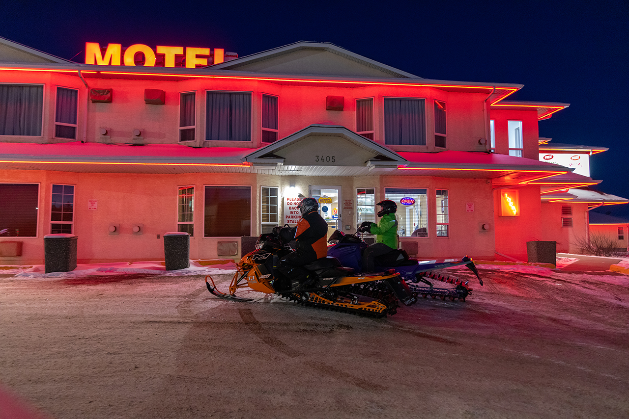 A Motel is lit up red with two snowmobilers parked in front of the building. 
