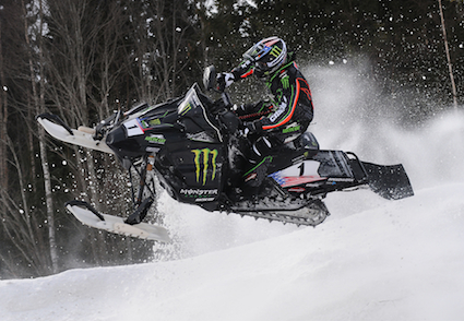 A man riding a snowmobile lifts off of the ground after taking a jump.