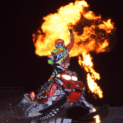 A snocross racer taking a jump and punching the sky with a fire ball behind him. 