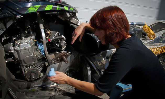 Krista, with short red hair and wearing a black shirt, cleans the inside of her snowmobile with a rag.