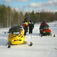 Yellow snowmobile in foreground with three snowmobilers and two sleds farther back, on a wide groomed trail edged by bush, on a sunny day