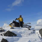 A rider in a yellow jacket sits on his snowmobile at the top of a snowy crest. A dramatic blue sky contrasts the snow and gray rocks.