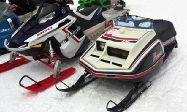 A vintage black, white and red Polaris snowmobile is parked next to a new black, white and red polaris snowmobile.