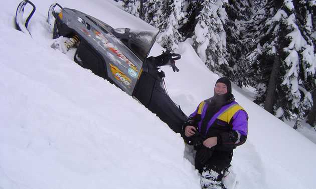 A snowmobile is stuck in deep snow on a 45-degree-angle hillside, with disgusted-looking rider standing by.
