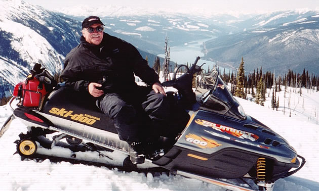 Smiling man relaxes on his Ski-Doo atop a snowy hill with a background of mountains