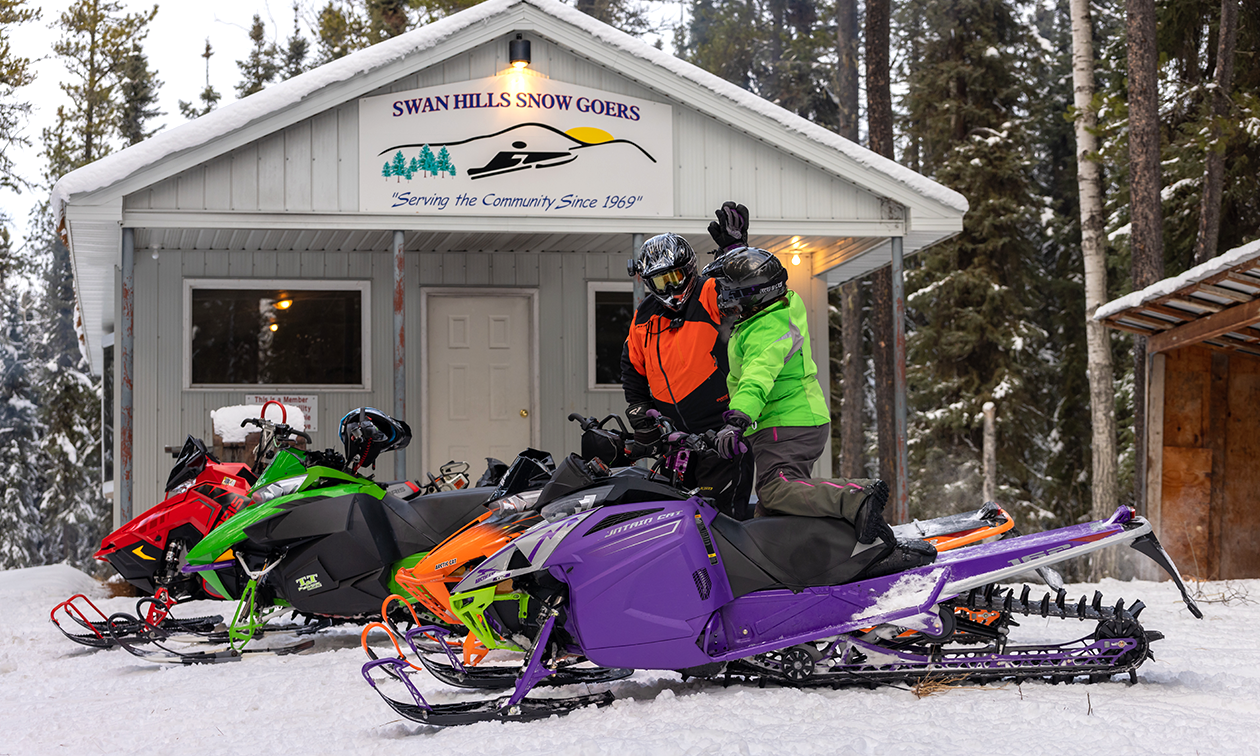 Two snowmobilers high five each other while standing upright on their snowmobiles in front of a Swan Hills Snow-Goers warm-up shelter