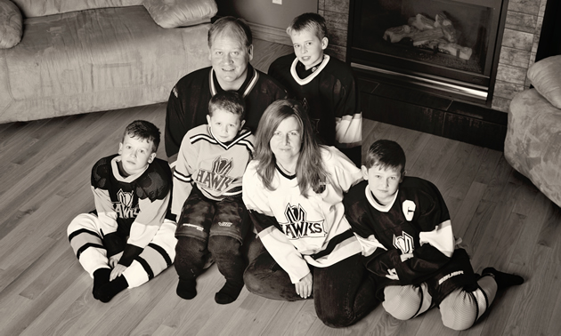Black and white photo of a man, woman and four young boys—all wearing hockey jerseys—sit on the floor in front of a fireplace