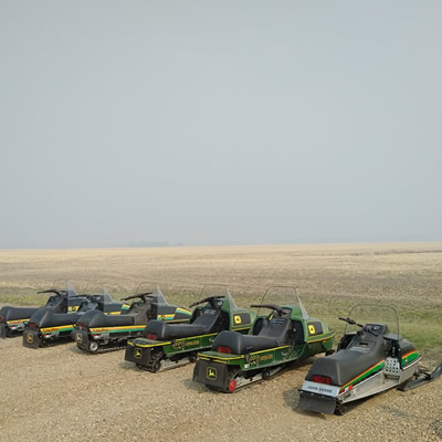 A row of John Deere snowmobiles out in a flat prairie field. 