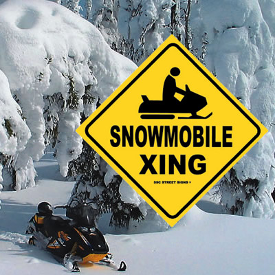 Snowmobile parked under snowy tree, with yellow 'Snowmobile Xing' sign. 