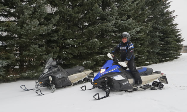 Snow-covered trail among evergreens has one riderless snowmobile and one with a male rider