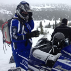A man dressed in a blue snowsuit and helmet looks at the camera while, behind him, two other riders observe the snowy backdrop.