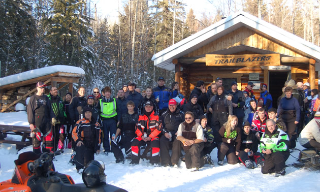 Crowd of 50 or more happy-looking people gather outside a warm-up cabin on a sunny winter day