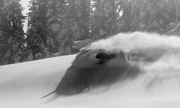 A black and white photo of a snowmobiler laying down a slash in the powder.  