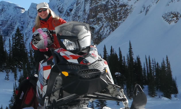 A blonde haired lady in red sitting on her snowmobile. 