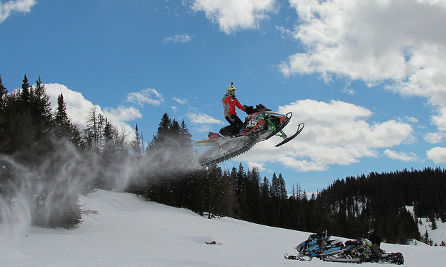 A plume of snowy powder trails behind a snowmobiler in the air.