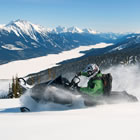 Photo of a guy riding a snowmobile fast across the snow on a ridge overlooking a large valley with mountains on either side. 