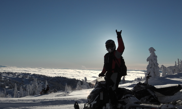 A silhouette of a woman standing on a snowmobile above the clouds. 