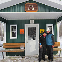 A man and a woman standing on the front step of a snowmobile cabin. 