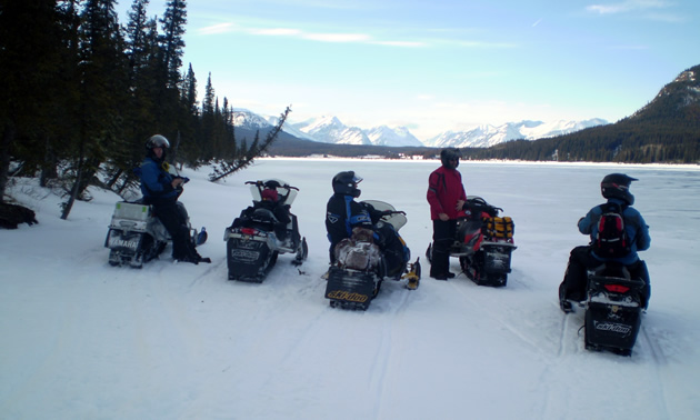 Five snowmobilers sitting on a lake looking out at the Rocky Mountains in the distance. 