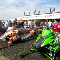 People milling around a green arctic cat and an orange and white arctic cat. 