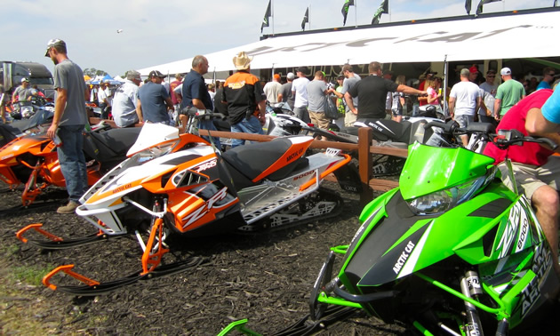 People milling around a green arctic cat and an orange and white arctic cat. 