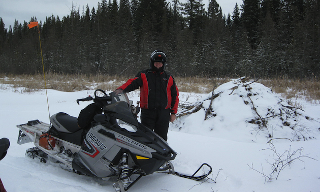 Man standing beside a snowmobile and a beaver lodge.
