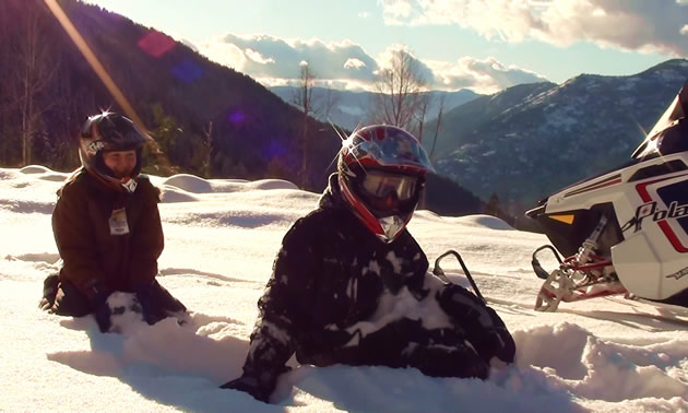 Photo of two young people sitting on the snow on top of a mountain.