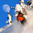 Photo of a guy hanging on a snowmobile as it launches over a cornice. Blue sky and a full moon is in the background. 