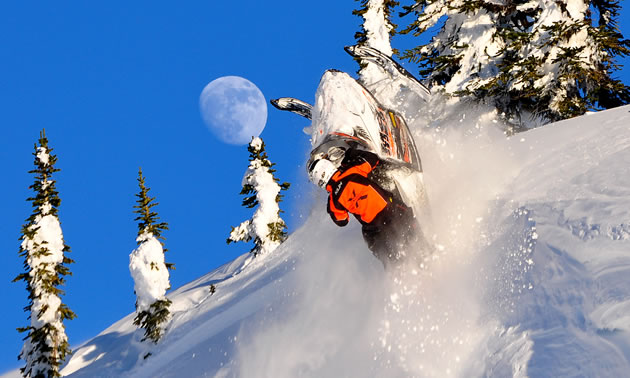 Photo of a guy hanging on a snowmobile as it launches over a cornice. Blue sky and a full moon is in the background. 