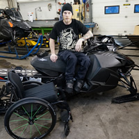 A young man sitting on a black sled with his wheelchair beside him. 