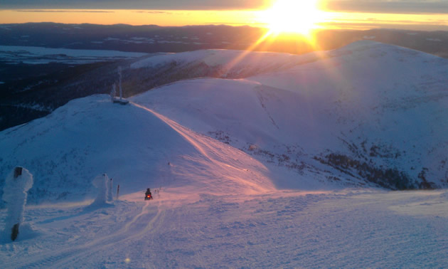 Photo of a person riding a snowmobile along a ridge with the sunset in the foreground.