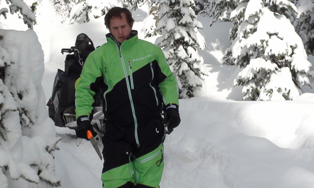 A man in a green Motorfist onesie holiding a saw. 