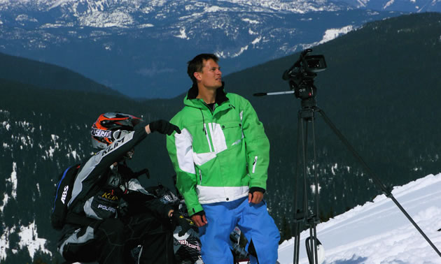 A man in a bright green jacket and a guy on a snowmobile looking to the mountains. 