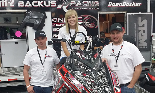A blonde haired woman on a sled with two men standing beside it. 